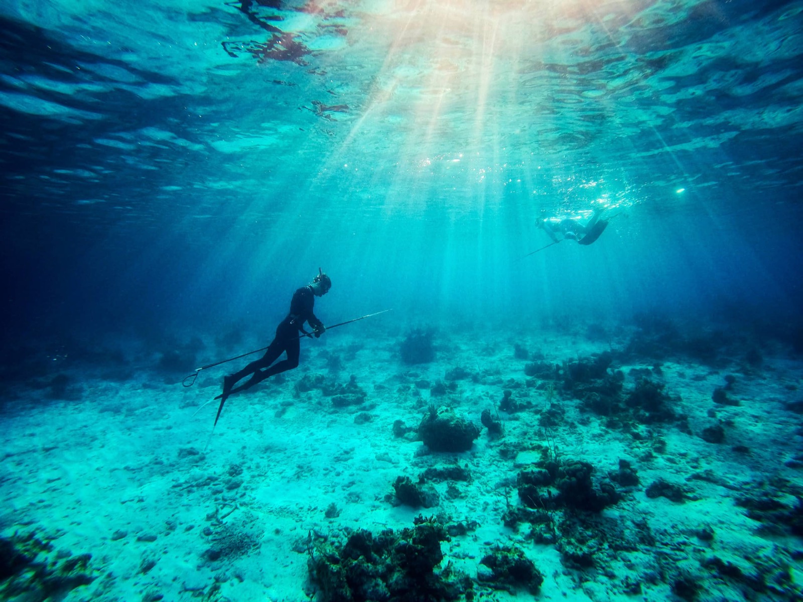A spearfisher gliding over sunlit coral, light rays pouring down from the surface