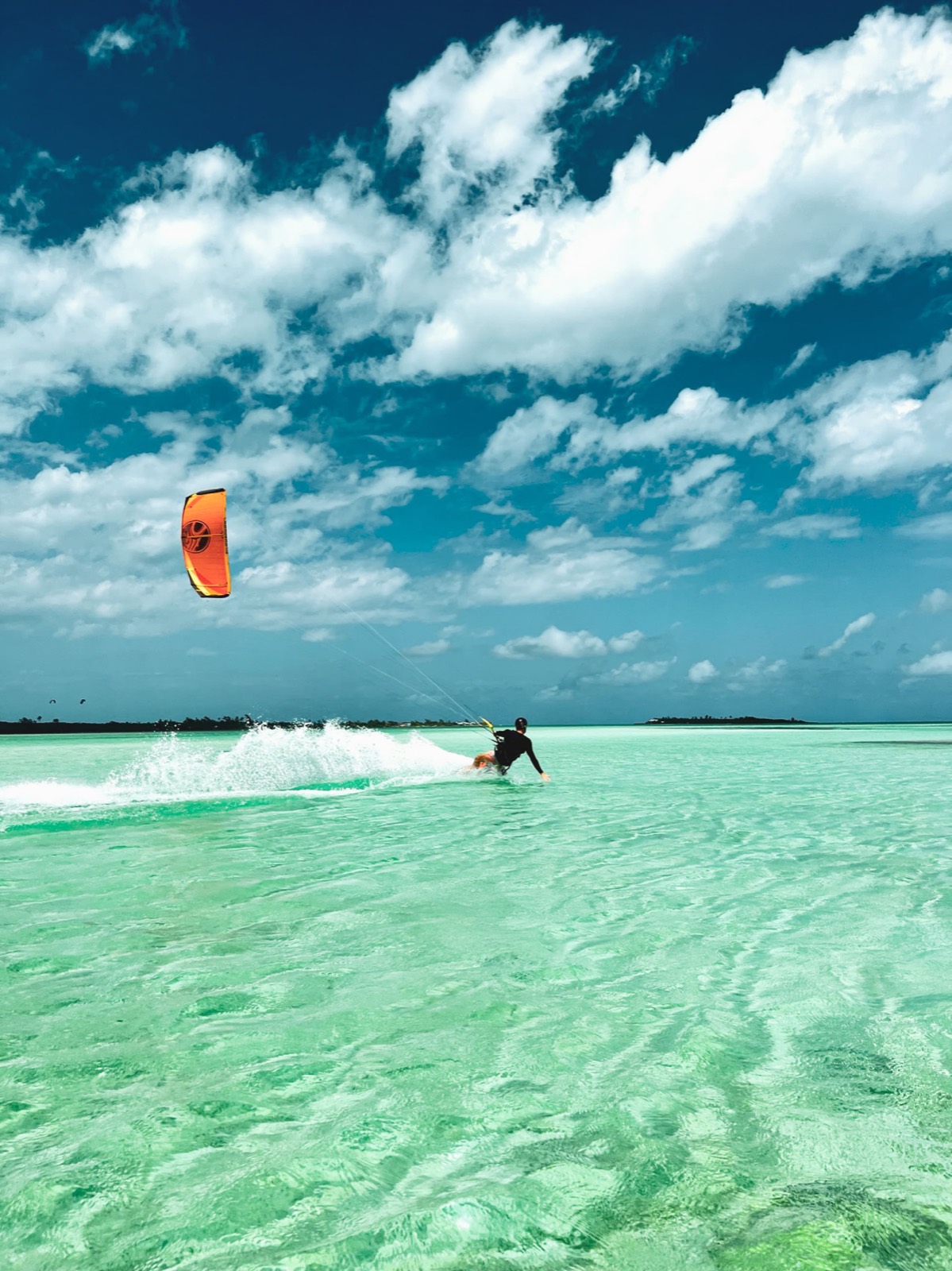 A kiteboarder carving across shallow turquoise flats, an orange kite against a deep blue sky