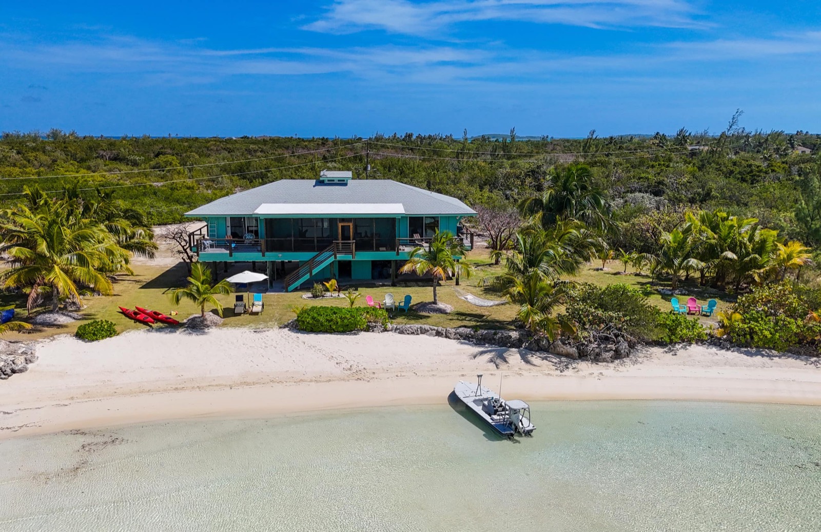 Aerial view of Villa Blue Hole from over the water: the teal-roofed house behind a secluded white-sand beach, a boat moored on the turquoise flats