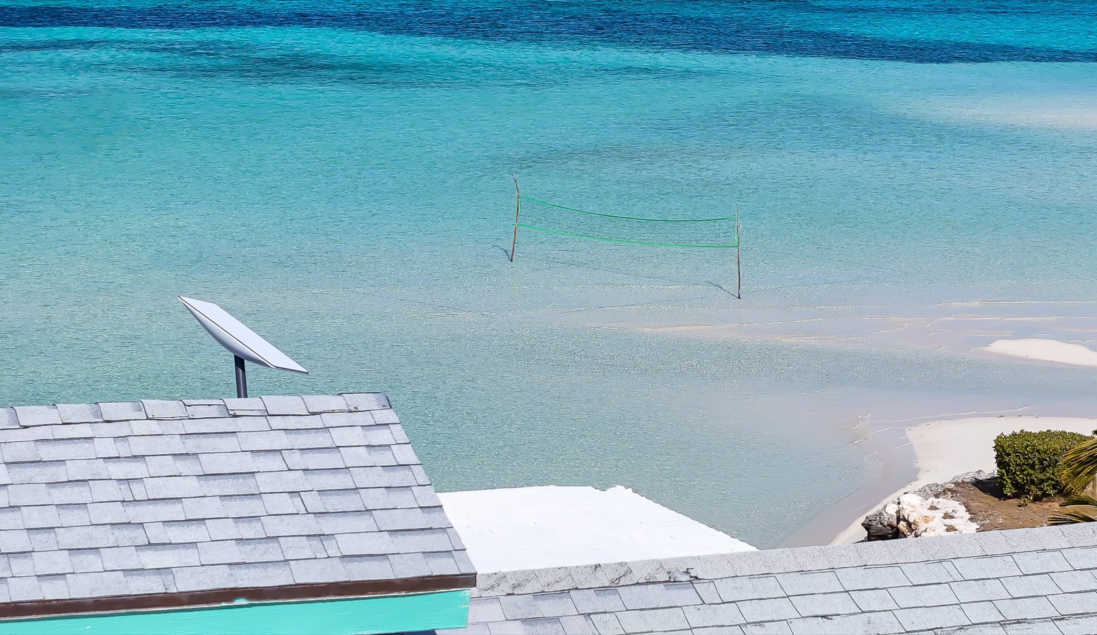 Rooftop view over the shingles and the Starlink dish onto turquoise flats with a beach-volleyball net standing in the shallows