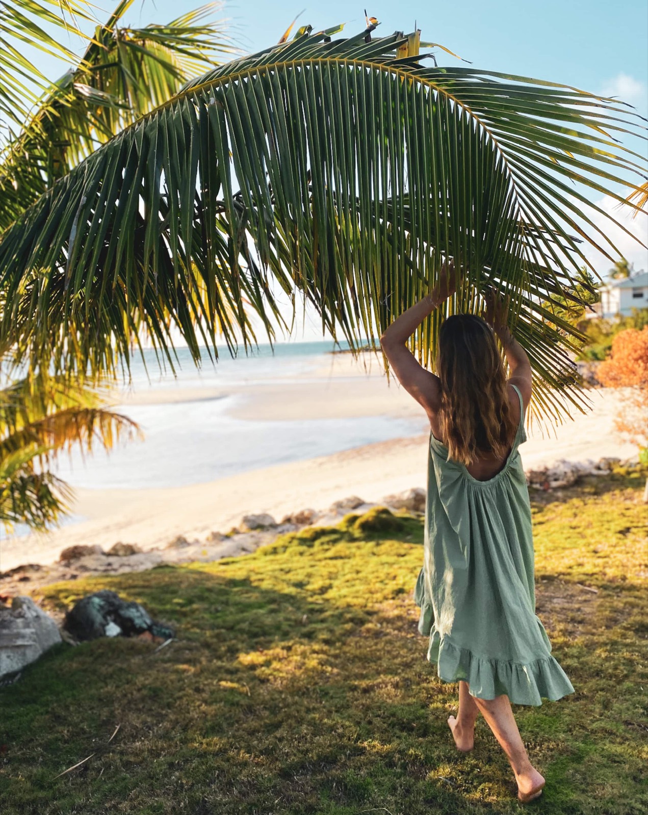 A woman in a sage-green dress walking from the lawn toward the beach in golden-hour light, lifting a palm frond