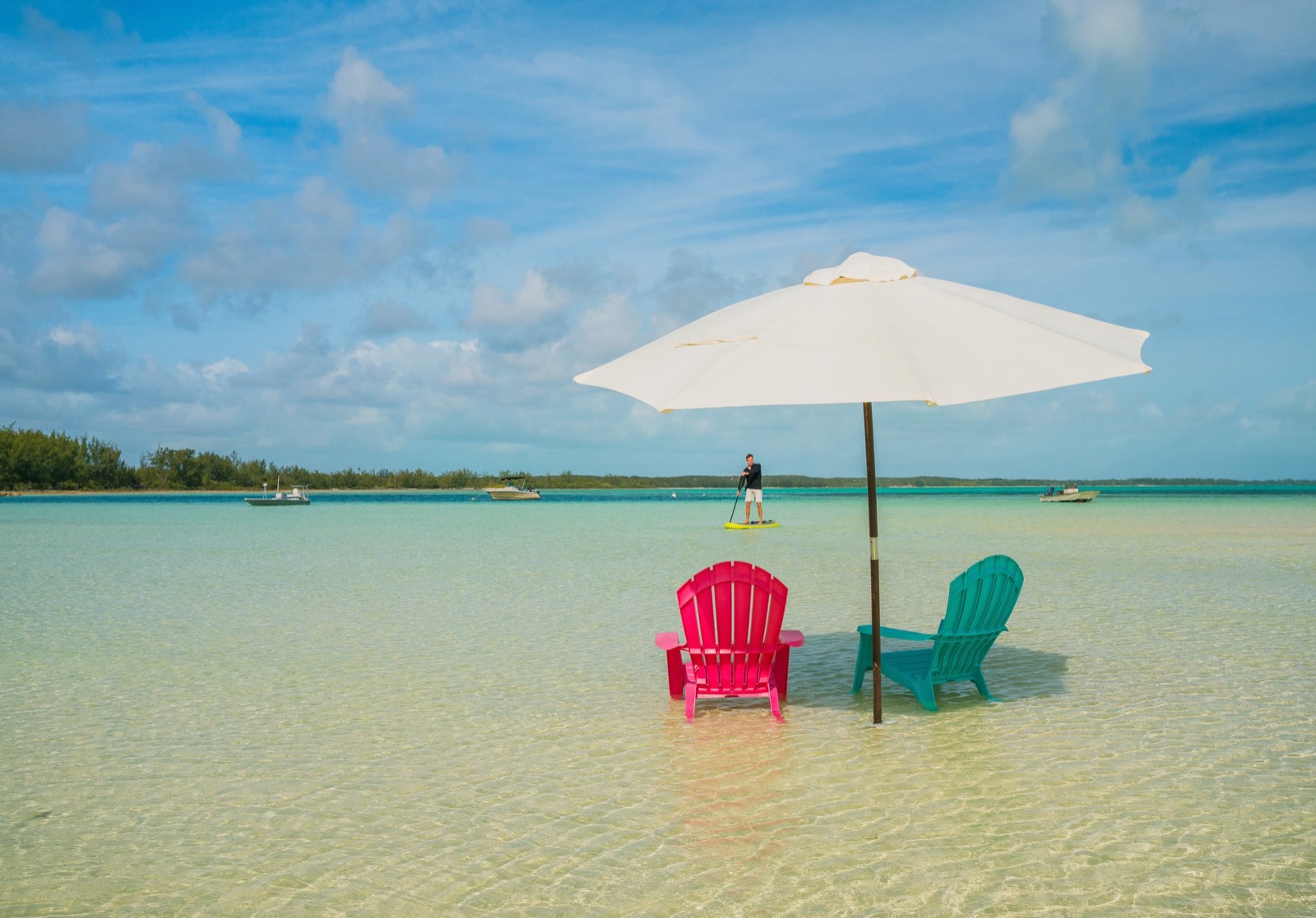 Two Adirondack chairs and a white umbrella standing in ankle-deep clear water on the flats, a paddleboarder in the distance